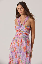 The woman is wearing a sleeveless, floral dress with a fitted bodice featuring a twisted front design and cut-out detail at the midriff. The dress has wide shoulder straps and a layered, flowing skirt with a pastel-colored floral pattern in shades of pink, orange, purple, and green.